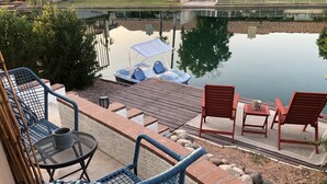 Un balcon avec des fauteuils en osier, une table avec une tasse, et une vue sur un lac avec un quai et un bateau.