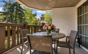 A round table with chairs, a vase with flowers, and two wine glasses on a balcony with a view of trees and a building.
