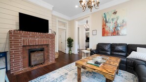 A living room with a brick fireplace, a black leather sofa, a wooden coffee table, and a large abstract painting on the wall. - 1920's Sunny Gem - Downtown Waco Home (Waco)