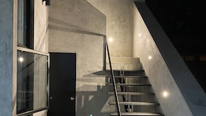 Una escalera con barandilla de metal que conduce a una puerta oscura, rodeada de paredes de hormigón y un ventanal.