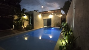 Una piscina con superficie de agua azul cristalina, rodeada por una terraza de concreto y un pequeño jardín con plantas en macetas.
