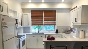 A modern kitchen with white cabinets, a stainless steel refrigerator, a built-in oven, a sink with a window above, and two black bar stools.
