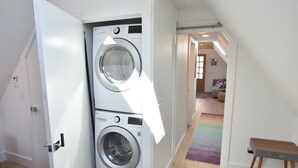 A laundry room with two stacked washing machines, a wooden bench, and a rainbow-colored rug. - 1 Min. Walk to Water/Town Deck and A/C! (Provincetown)