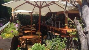 An outdoor seating area with wooden tables and chairs, surrounded by greenery and a pergola with a white umbrella. - 1 Min. Walk to Water/Town Deck and A/C! (Provincetown)