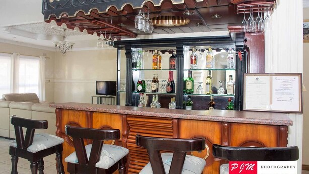 Eine Bar mit Holztresen und drei Stühlen. Hinter dem Tresen befinden sich Regale mit Flaschen und Gläsern.