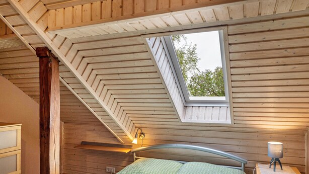 Een slaapkamer met een schuine kap, dakraam, een bed met groene beddengoed, een bedlampje en een houten nachtkastje.