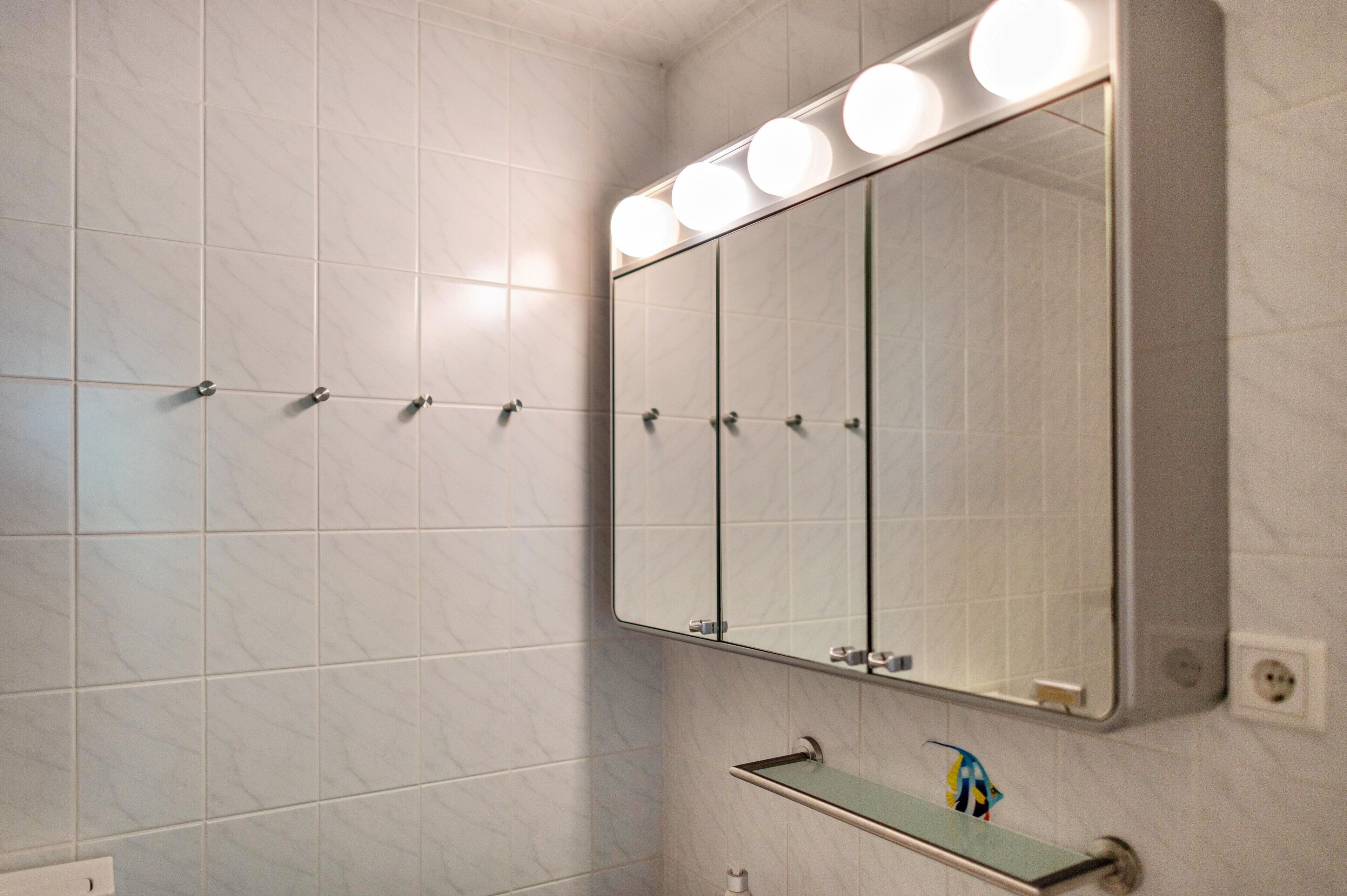 Un baño con baldosas de mármol blanco, un espejo grande con luces y un estante con una toalla.