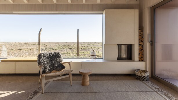 A modern interior with a large window offering a view of the desert landscape. There is a seating area with a bench and a chair, a small table, and a fireplace. - Radar by Bloom Stays - sleeps 4 on nature reserve (Dungeness)