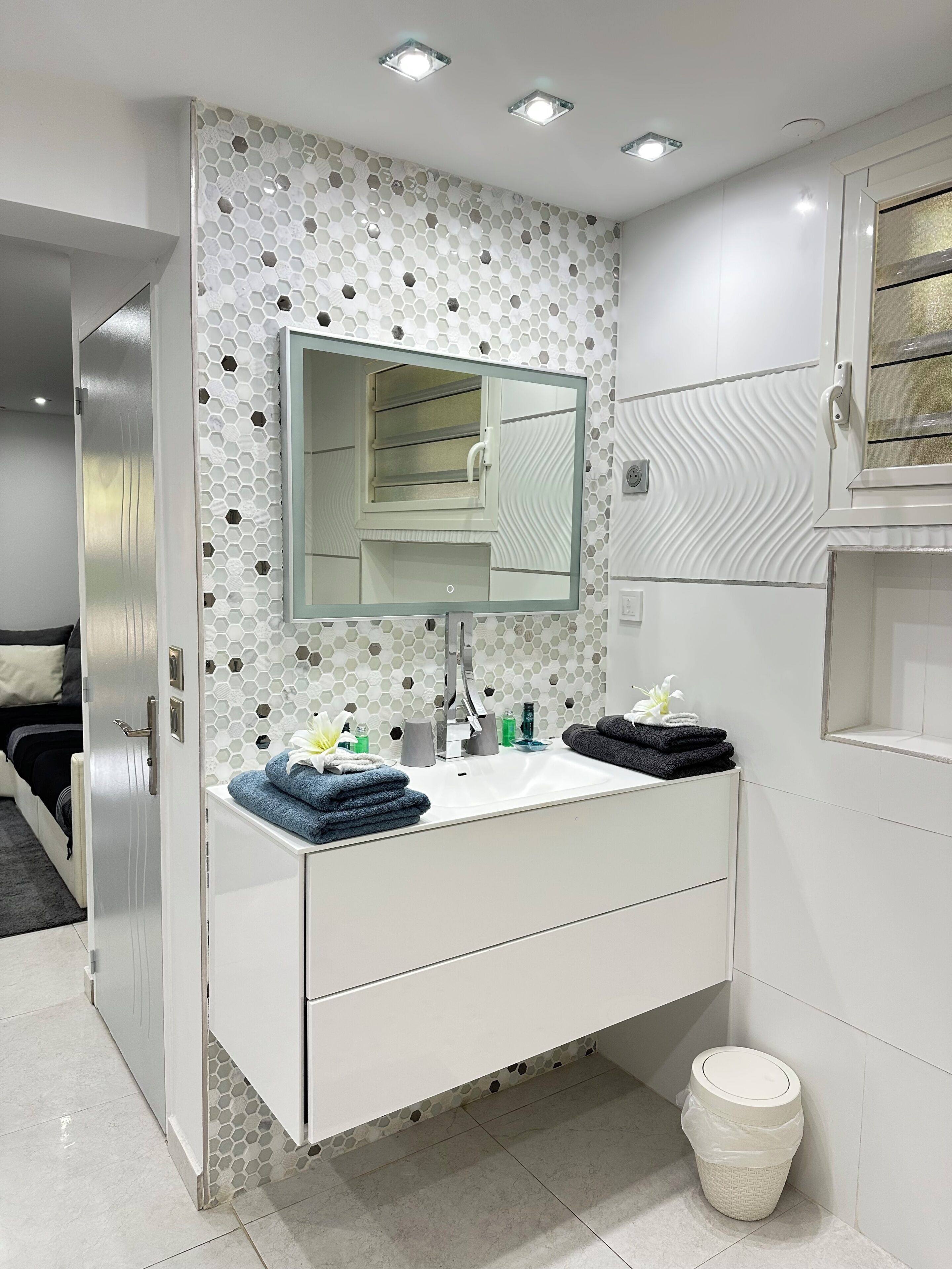 Une salle de bain moderne avec un meuble-lavabo blanc, un grand miroir, un mur orné d’un motif hexagonal et une poubelle blanche.
