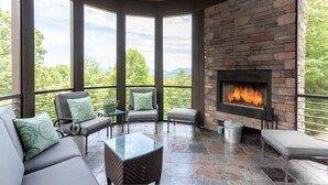 A covered outdoor seating area with a stone fireplace, large windows, and a view of greenery. - Yourcation® Awaits in Majestic Mountain Home! (Fairview)
