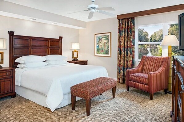 A hotel room with a large bed, a wooden headboard, a bench, a striped armchair, a ceiling fan, a window with curtains, and a painting on the wall. - Beautiful updated Condo in World Class Resort Near Disney (Orlando)