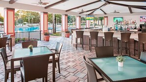 An outdoor bar area with wicker furniture, a pool in the background, and a bar stocked with bottles. - Beautiful updated Condo in World Class Resort Near Disney (Orlando)