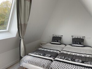 Ein kleines, modernes Schlafzimmer mit schrägem Dach, zwei Betten mit gemusterten Bettwäsche, einem Nachttisch und einem Fenster mit Vorhängen.