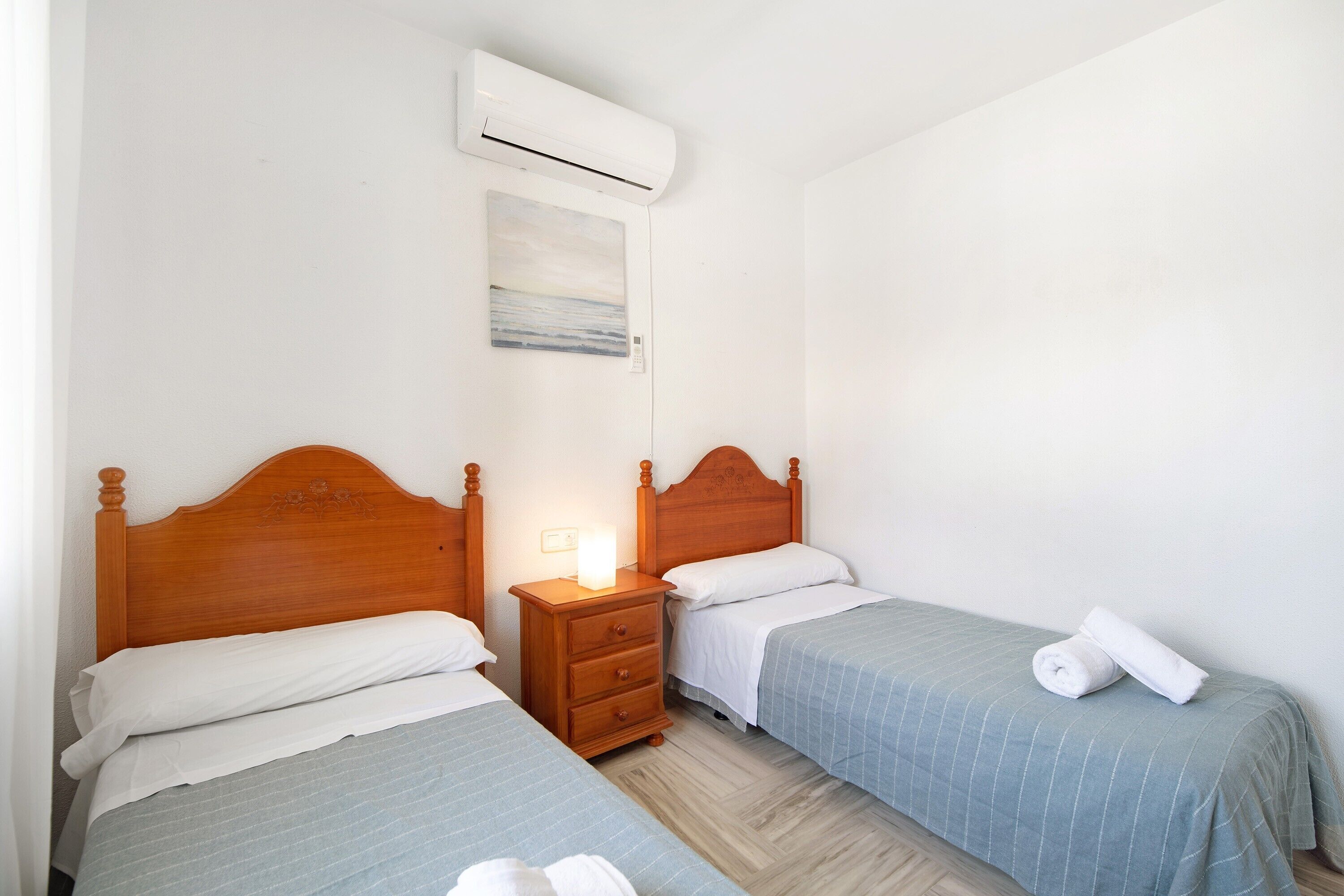 Habitación de hotel con dos camas, cabecero de madera, mesita de noche con lámpara y equipo de aire acondicionado.