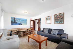 Olohuone, jossa on sohva, sohvapöytä, ruokapöytä tuoleineen sekä taulu, jossa on kuvattuna merimaisema.