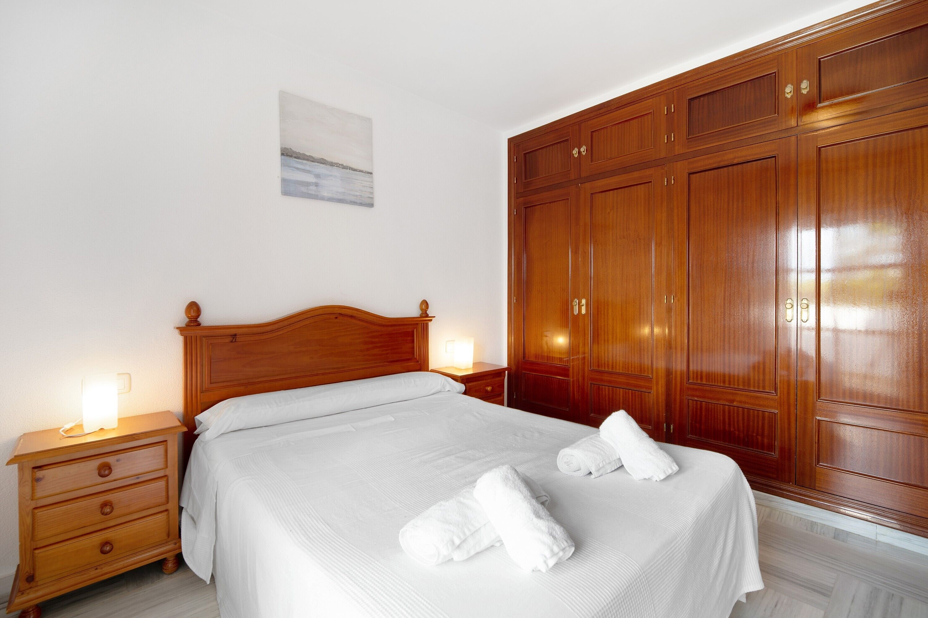 A neatly made bed with white linens and towels, a wooden nightstand with a lamp, and a large wooden wardrobe.