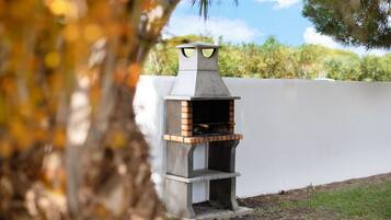 Een buitenbarbecue van beton met een schoorsteen, voor een witte muur en omringd door gras en bomen.