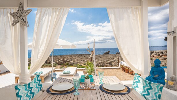 Un área de estar al aire libre techada con vista al mar, mesas de madera, cortinas blancas y una estatua de Buda.