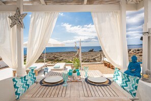 Un área de estar al aire libre techada con vista al mar, mesas de madera, cortinas blancas y una estatua de Buda.