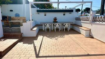 Un patio con mesa y sillas, un pequeño jardín y una pared blanca con azulejos decorativos.
