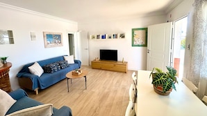 Een woonkamer met een bank, een salontafel, een tv op een houten standaard, een boekenkast en een deur.