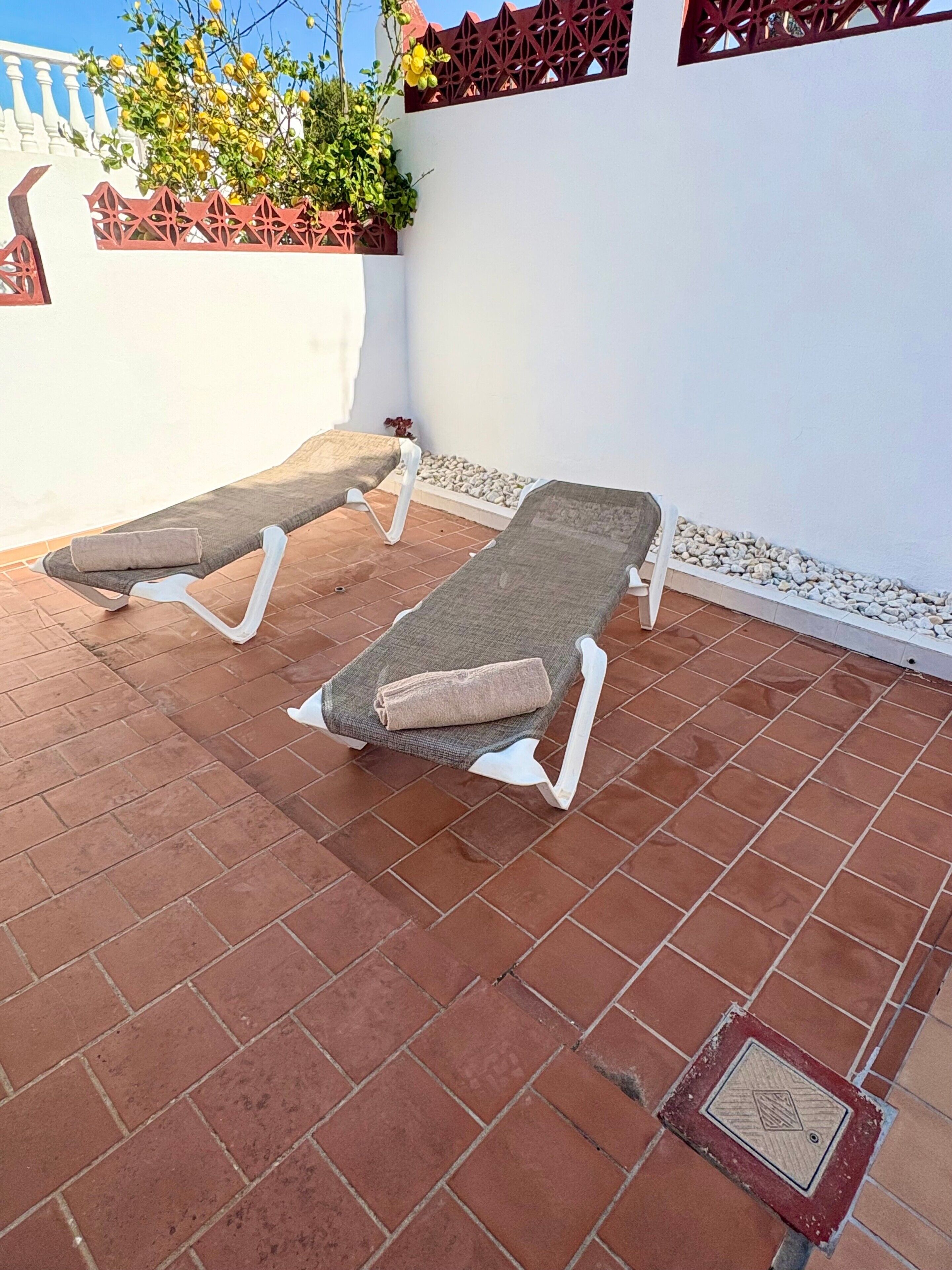 Dos tumbonas en un patio con suelo de baldosas, una pared blanca y una valla decorativa al fondo.