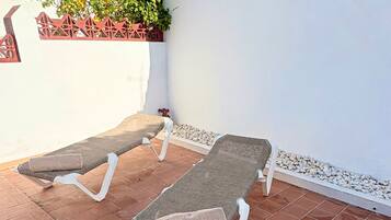 Dos tumbonas en un patio con suelo de baldosas, una pared blanca y una valla decorativa al fondo.