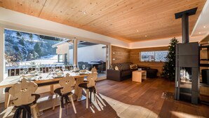 Un intérieur moderne avec un plafond en bois, de grandes fenêtres, un coin repas avec des chaises et une table dressée pour quatre personnes, un canapé et une cheminée.