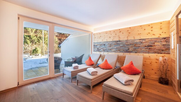 Un moderno spa con puerta de vidrio que da al exterior, suelo de madera y un rincón de descanso con toallas y cojines.