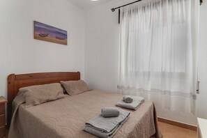 Une chambre à coucher avec une tête de lit en bois, un lit recouvert d’une courtepointe beige, deux oreillers, une table de chevet, une serviette enroulée et un tableau accroché au mur.