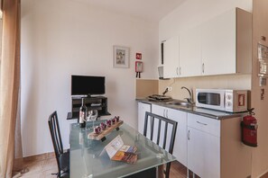A compact kitchen with a microwave, toaster, and sink. There is a glass table with a wine bottle and glasses, and a chair. A television is mounted on the wall.