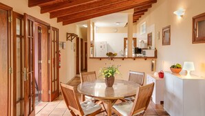 A dining area with a round table, wooden chairs, a vase with flowers, and a kitchen in the background. - Vacation Home "Elvira Hermigua" with Sea View, Mountain View, Terraces & Garden (Hermigua)