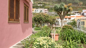 A pink building with wooden shutters, a garden with yellow flowers, and a hillside in the background. - Vacation Home "Elvira Hermigua" with Sea View, Mountain View, Terraces & Garden (Hermigua)