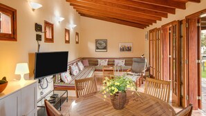 A cozy living area with a wooden ceiling, a television, a dining table with chairs, a sofa, and a window with wooden shutters. - Vacation Home "Elvira Hermigua" with Sea View, Mountain View, Terraces & Garden (Hermigua)