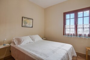 A bedroom with a bed, a nightstand, a lamp, a window with curtains, and a poster on the wall. - Vacation Home "Elvira Hermigua" with Sea View, Mountain View, Terraces & Garden (Hermigua)