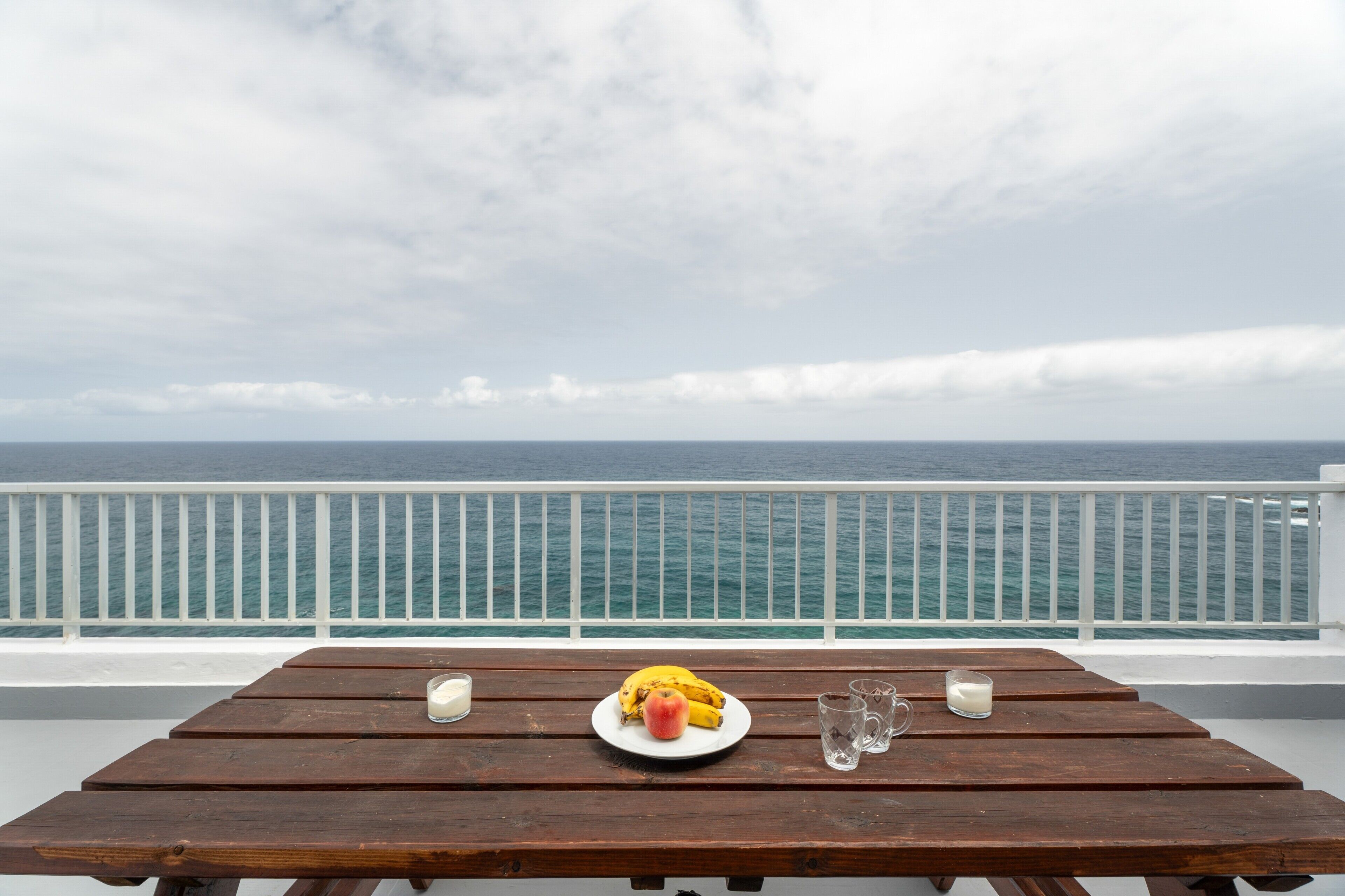 Una mesa de madera con un plato de comida, un vaso de leche y un plátano en un balcón con vista al mar.