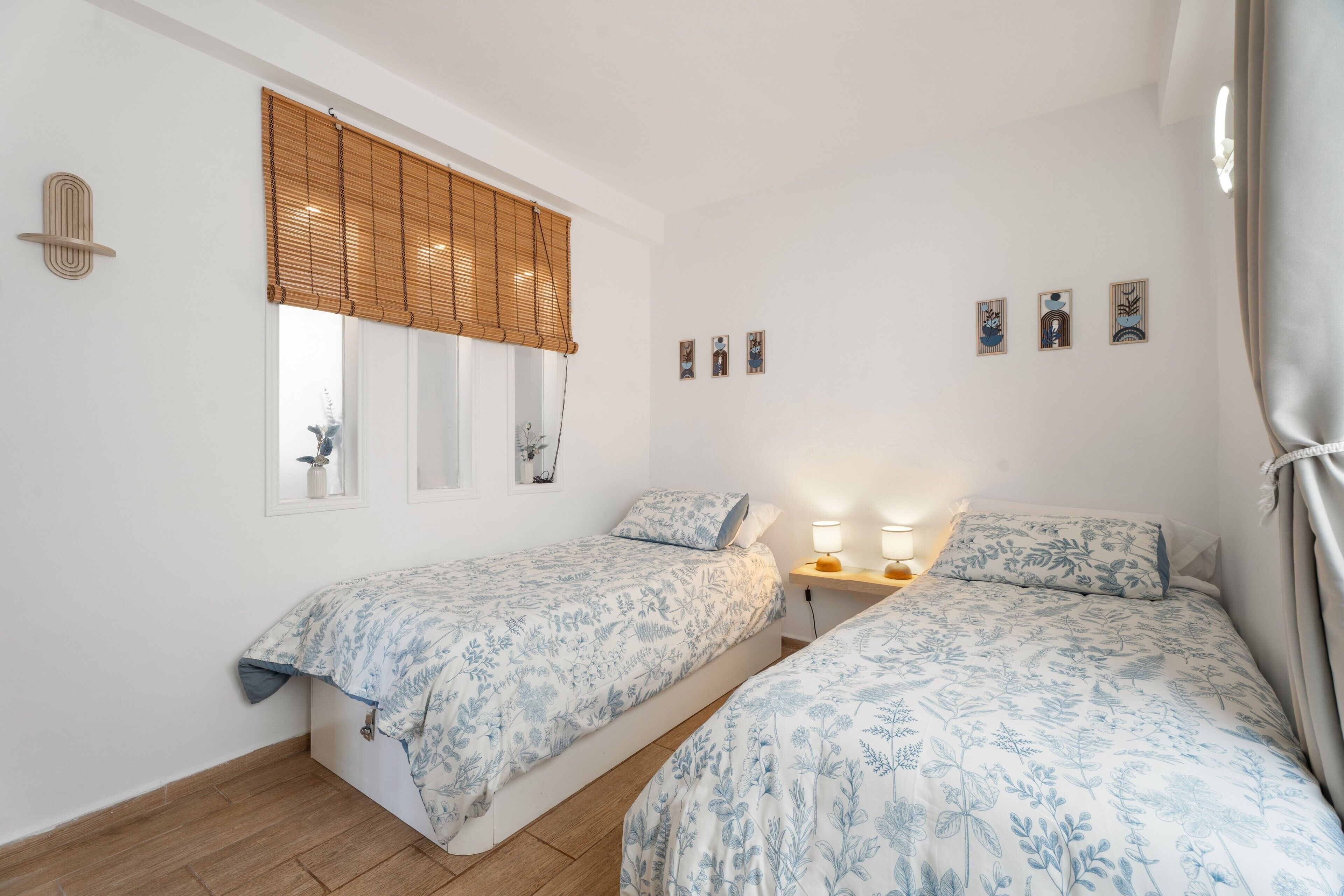 Un dormitorio con dos camas, una ventana con persianas, una mesita de noche con lámparas y una pared decorada con cuadros.