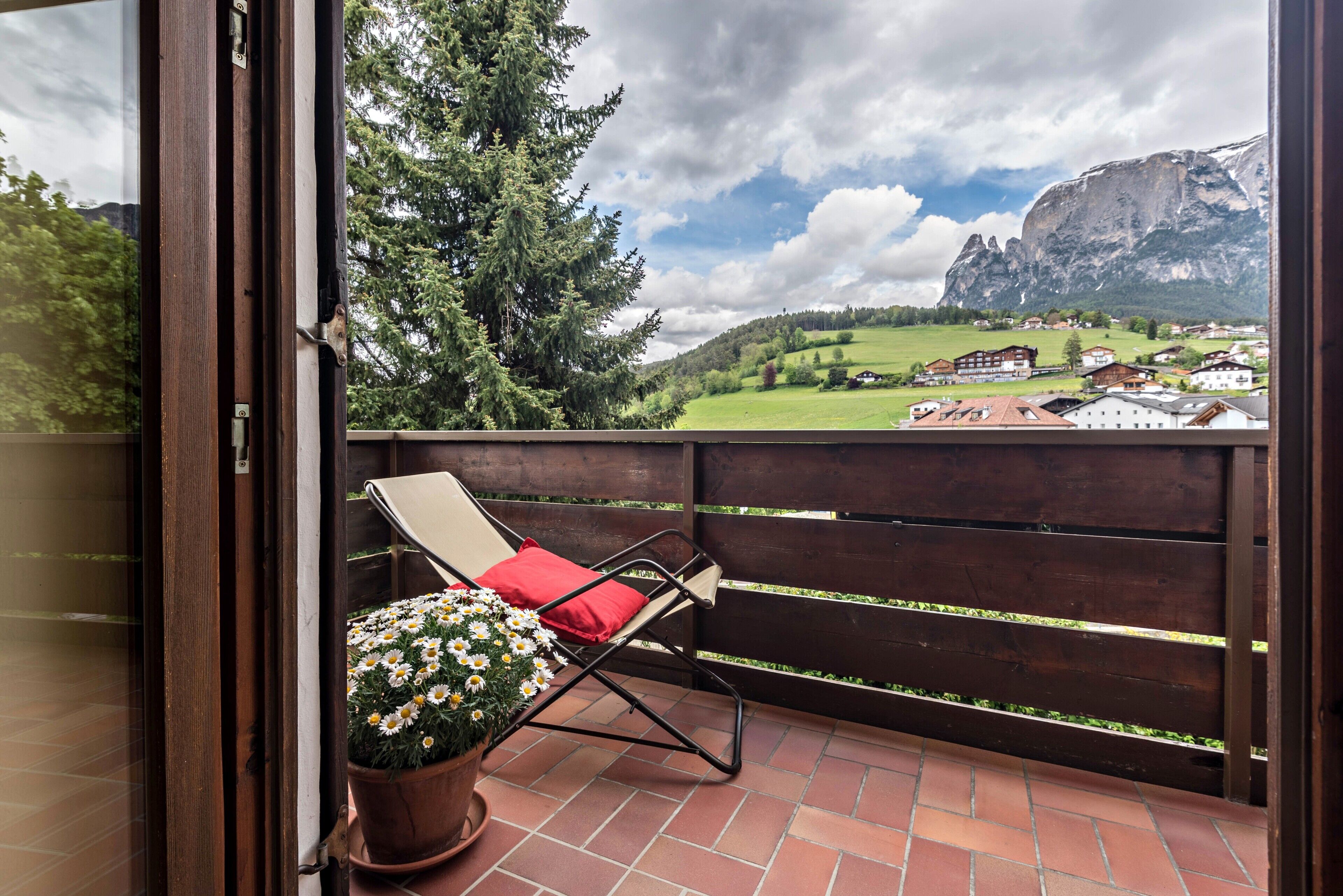 Un balcone con una sedia e una pianta in vaso, affacciato su un paesaggio montuoso e un villaggio.