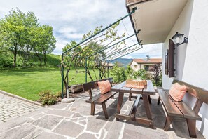 Une terrasse avec un sol en pierre, des bancs en bois, une table et une pergola couverte de plantes grimpantes.