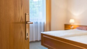 Uma porta de madeira com uma fechadura, uma cama com colchão branco e uma mesa de cabeceira com abajur.
