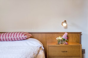 A neatly made bed with a plaid pillow, a bedside table with a vase of flowers, and a wall-mounted light fixture.