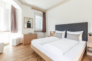 A modern hotel room with a large bed, a wooden floor, a small desk, a mirror, and a window with curtains. - Villa Martiner Apartments Francesco with Mountain View, Balcony & Wi-Fi (Ortisei)