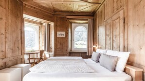 Ein Zimmer aus Holz mit einem Bett, zwei Fenstern, einem Sessel und einem Tisch.
