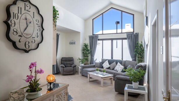 Ein geräumiges Wohnzimmer mit einer großen Uhr, einer Sofagarnitur, einem Couchtisch und Blick durch ein großes Fenster.