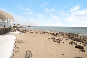 A beach with a building on the left, rocky shoreline, and clear sky.