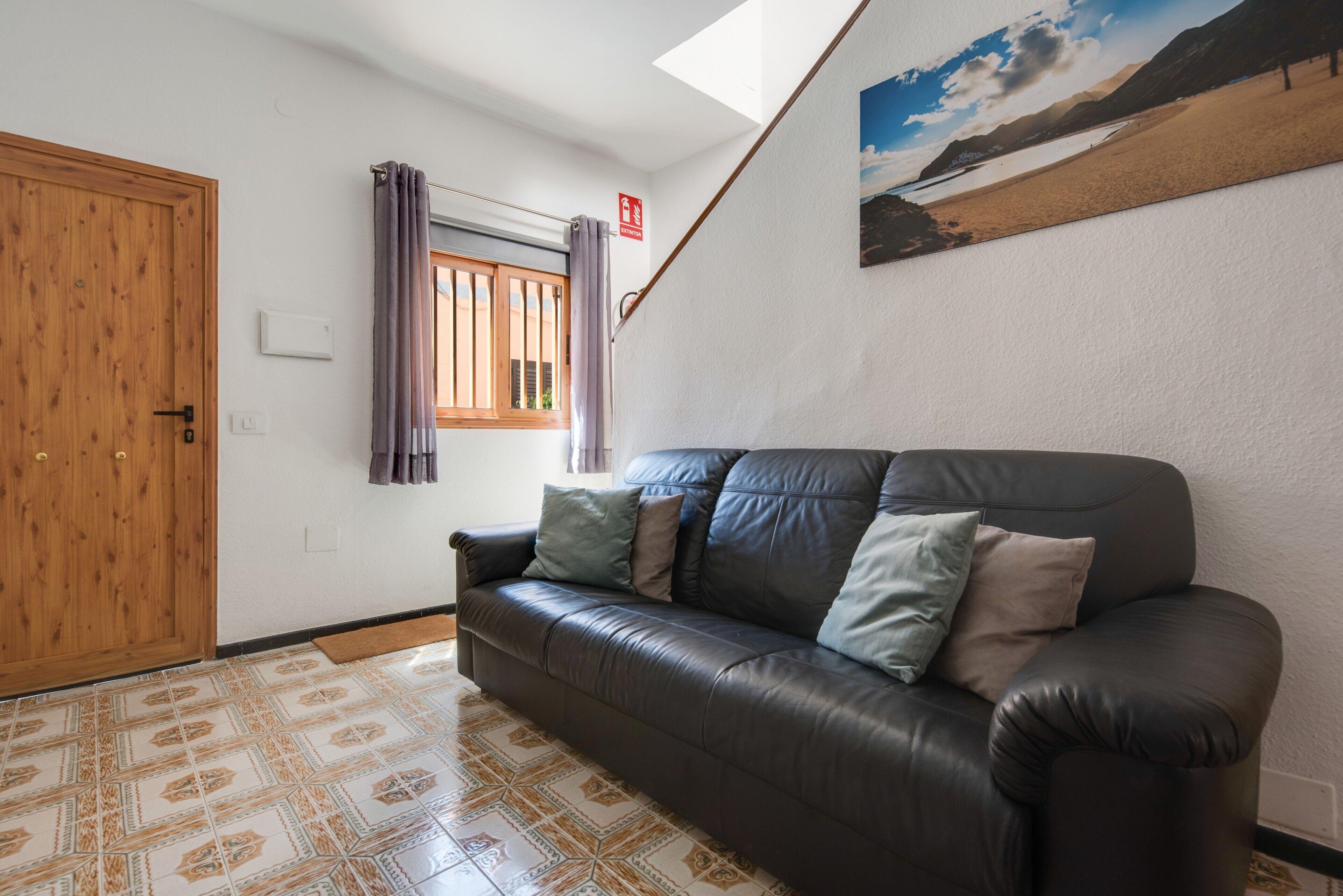 Quarto com sofá de couro escuro, ladrilhos estampados, porta de madeira, janela com cortinas e um quadro na parede.