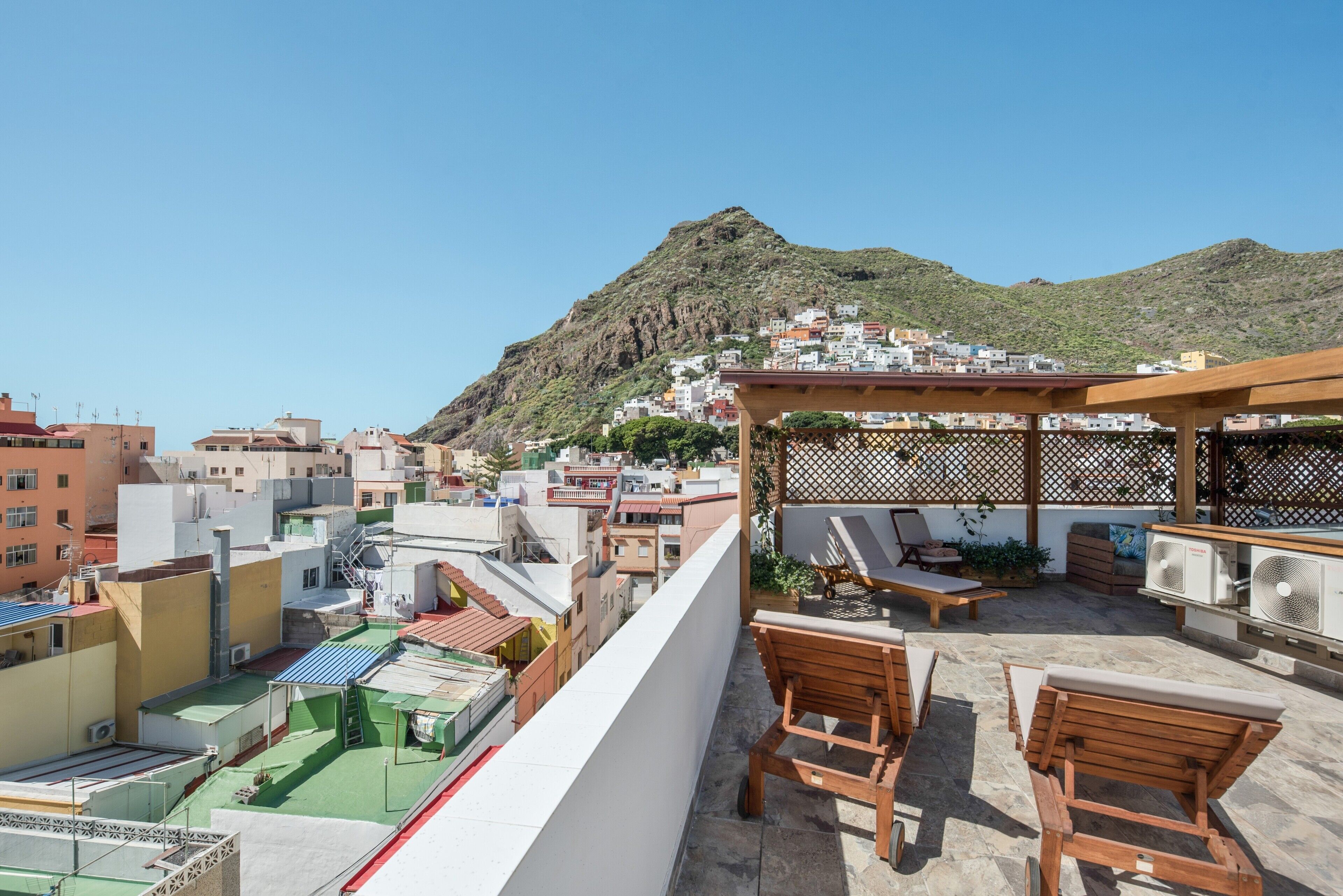 Une terrasse sur le toit, avec des chaises en bois, une pergola et une vue sur une ville située sur une colline.