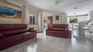 Una sala de estar con dos sofás de cuero, una mesa de comedor con sillas, una zona de cocina y un ventanal con cortinas.
