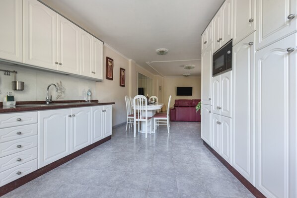A kitchen with white cabinets, a dining table with chairs, a red sofa, and a flat-screen TV mounted on the wall.