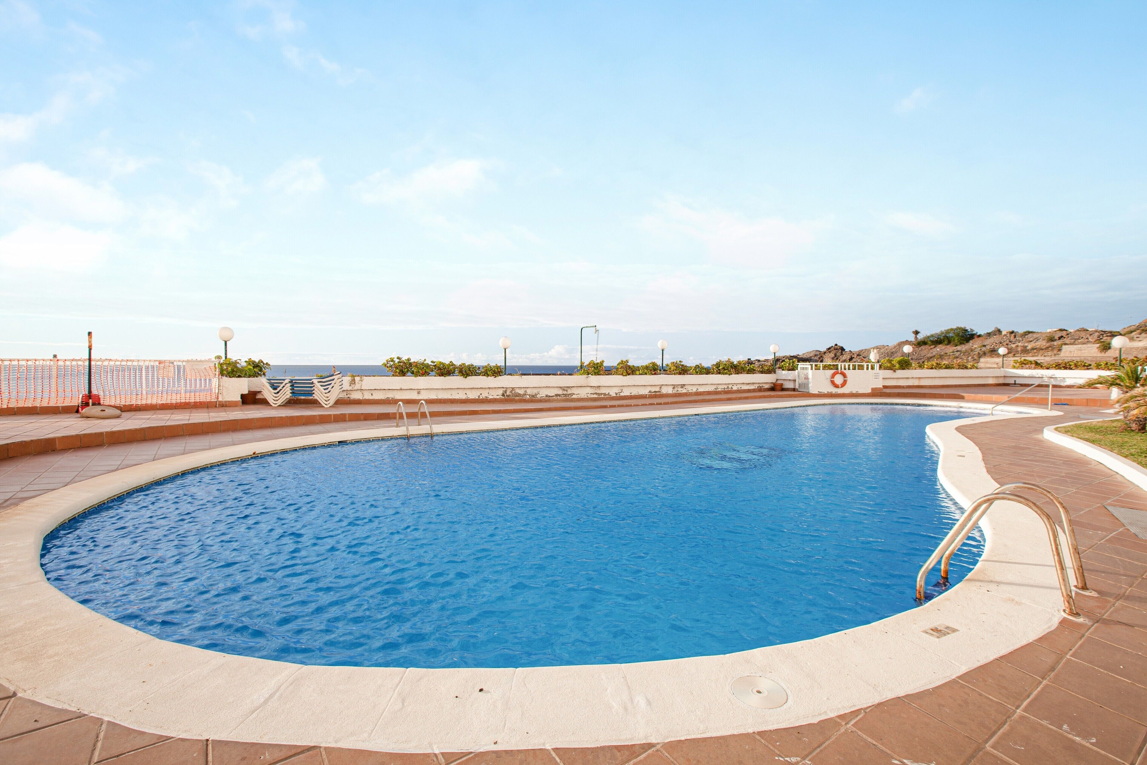 A swimming pool with a curved edge, a tiled deck, and a view of the sea in the background.
